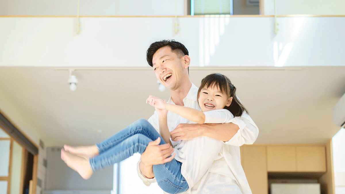 passiv energieのWEBコラム用イメージ写真。吹き抜けから自然光が優しく降り注ぐ明るい木造住宅の室内で、父親が小さな娘を笑顔で抱きかかえ、楽しそうにスキンシップをとって遊んでいる微笑ましい日常風景。高気密・高断熱住宅の快適性や、家族の健康を守る24時間計画換気をテーマにしたコラム記事向けの挿入画像