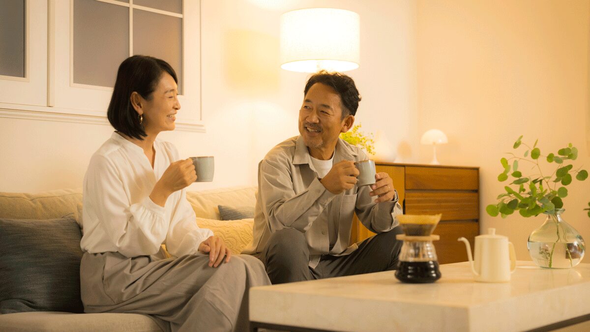 passiv energieのWEBコラム用イメージ写真。温かみのある間接照明が灯る落ち着いた雰囲気のリビングルームで、ソファに座りながらマグカップを手に笑顔で語り合うミドルシニア・シニア世代の夫婦。手前のローテーブルにはコーヒーカラフェやポットが置かれた、セカンドライフの快適な住環境づくりや、年中温度差のない健康的な家づくりをイメージさせる挿入画像